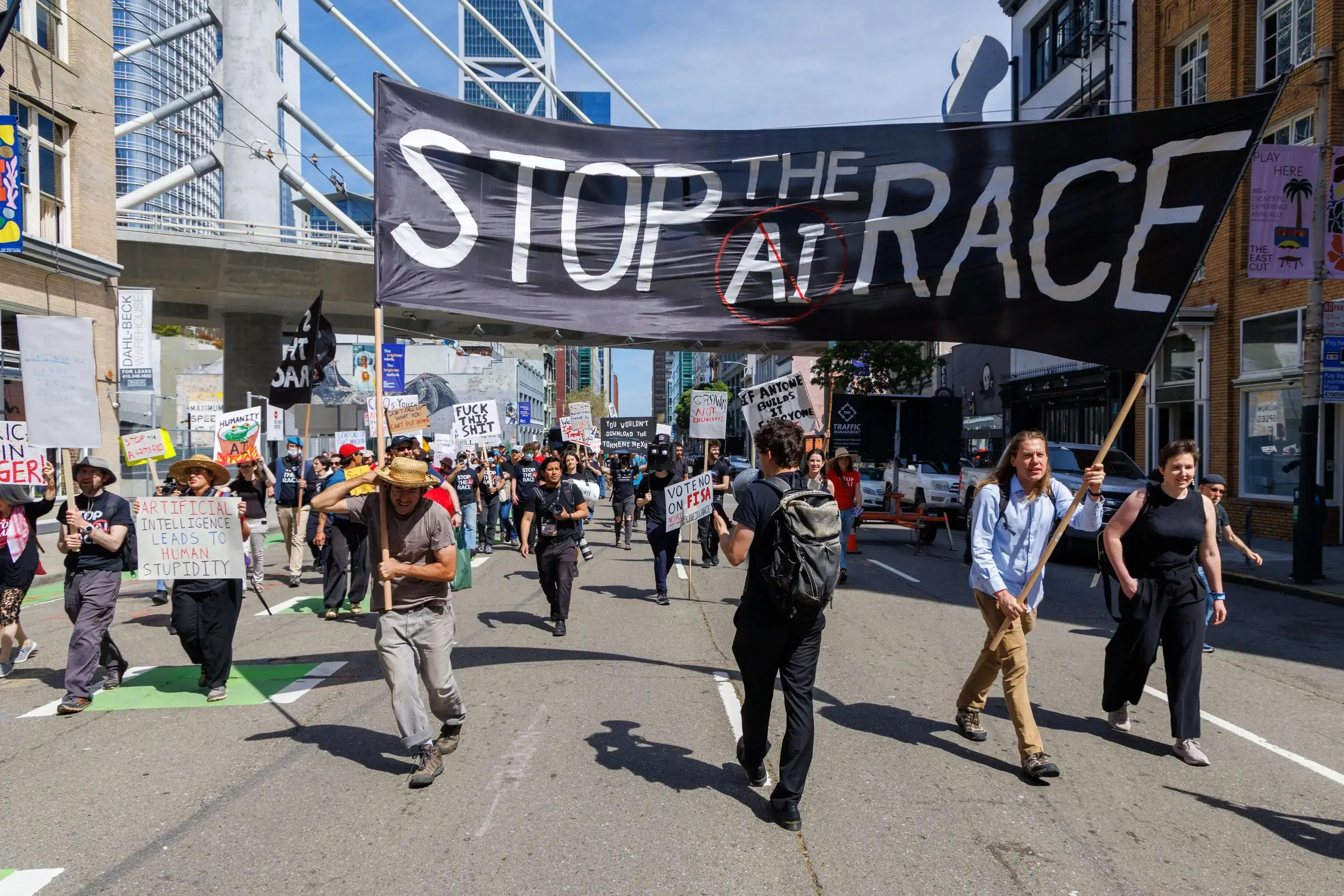 2026 年 3 月，针对美国 AI 竞赛的小型游行。组织者的口号是，希望 xAI、Anthropic 和 OpenAI 等 AI 巨头：如果其他人承诺停止，自己也停止竞赛。然而，AI 竞赛是一个全球正义问题，而不是一个单纯的技术安全问题。这些巨头本身就是资本。它们一方面向公众宣言，AI 可能会威胁人类，一方面劝说政府支持它们这样的企业开发 AI，因为只有它们才能确保 AI 的安全，但有的人却指出，这不过是为了垄断以盈利。像 Anthropic 这样的资本竟然展现出对安全如此魔怔的关心，还宣言在其“宪法”的指导下开发 AI，属实令人怀疑。